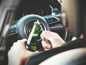 Person opening a beer bottle while sitting behind the wheel of a car, symbolizing the dangers of impaired driving and the need for a Drunk Driving Accident Attorney.